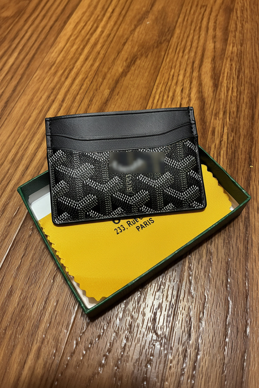A black monogram card holder wallet resting on its green and yellow branded presentation box, placed on a wooden table.